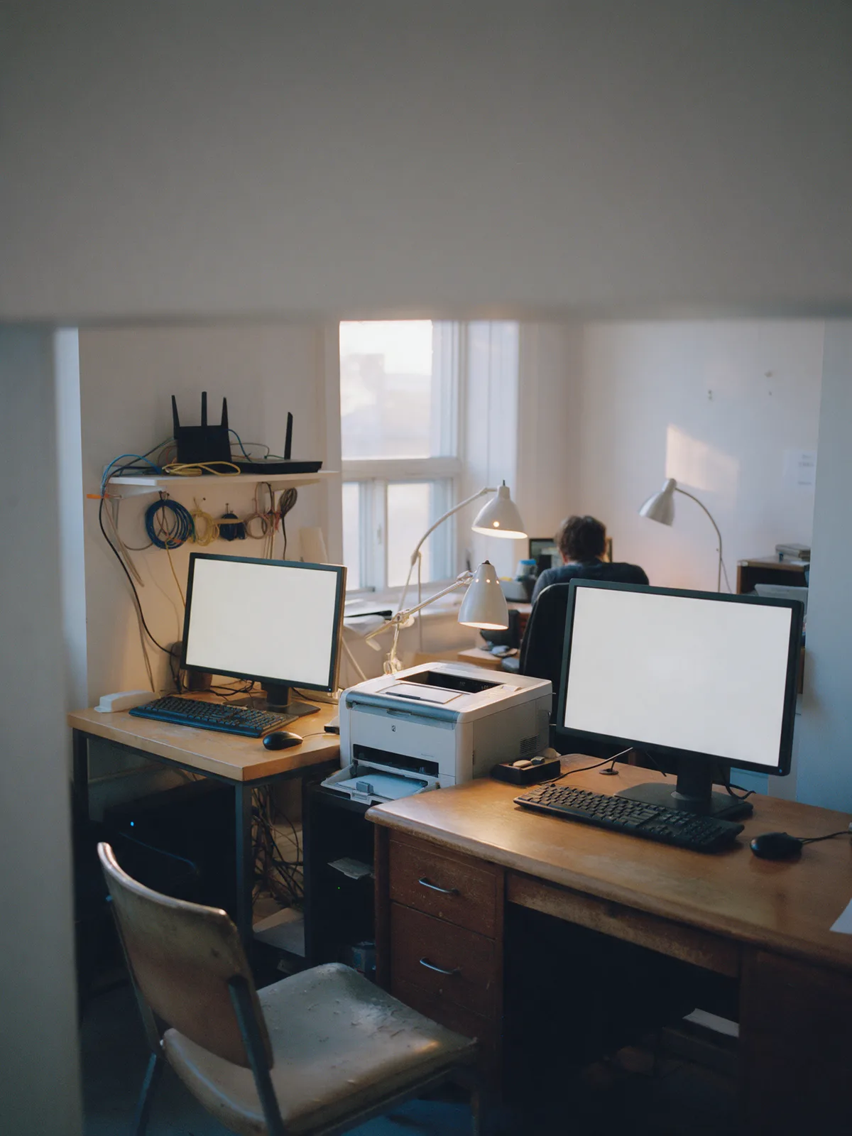 Un petit bureau avec deux postes de travail, un routeur et une imprimante, lumière de fin d’après-midi.